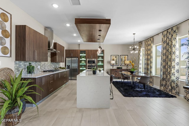 kitchen featuring tasteful backsplash, wall chimney exhaust hood, stainless steel appliances, a chandelier, and hanging light fixtures