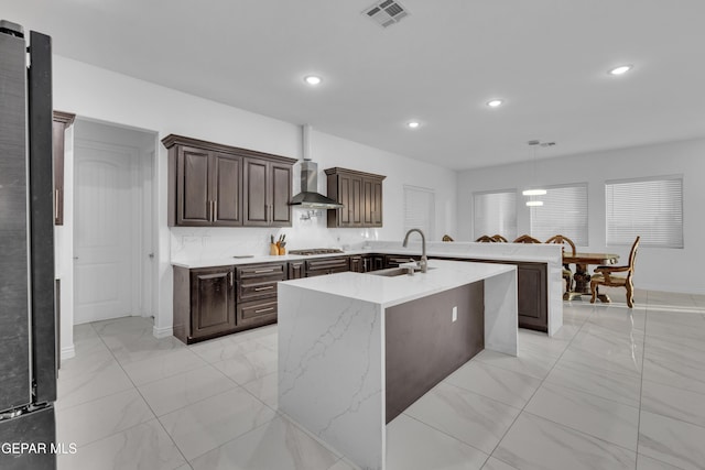 kitchen with wall chimney exhaust hood, sink, tasteful backsplash, hanging light fixtures, and an island with sink