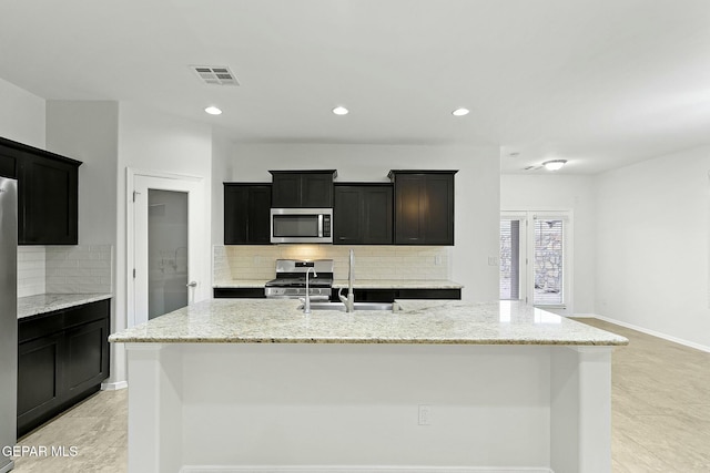 kitchen featuring a center island with sink, sink, decorative backsplash, light stone countertops, and stainless steel appliances