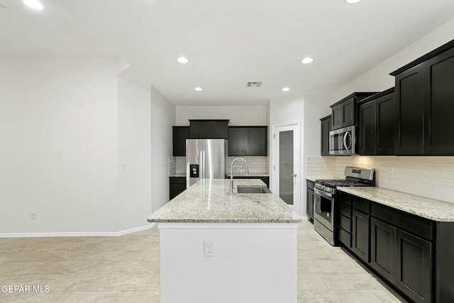 kitchen featuring light stone countertops, sink, stainless steel appliances, backsplash, and a center island with sink