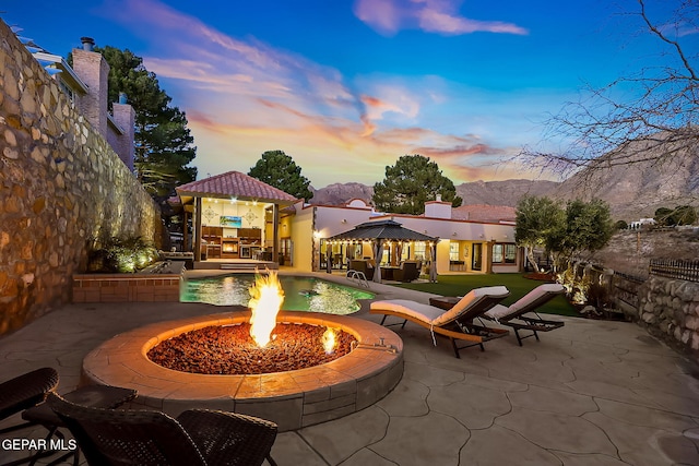 pool at dusk with an outdoor fire pit, a patio area, a gazebo, and a mountain view