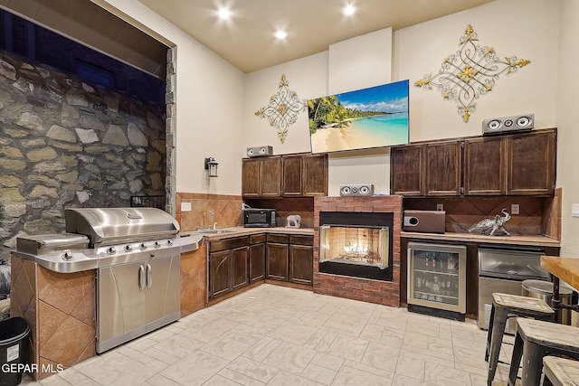 kitchen featuring a brick fireplace, decorative backsplash, wine cooler, stainless steel counters, and dark brown cabinetry