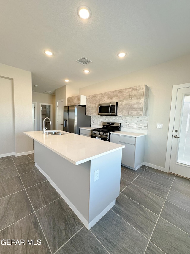 kitchen with sink, an island with sink, decorative backsplash, dark tile patterned flooring, and appliances with stainless steel finishes