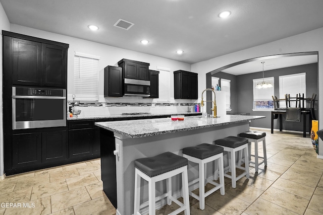 kitchen featuring a center island with sink, stainless steel oven, light stone countertops, and hanging light fixtures