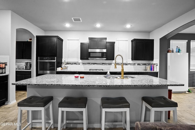 kitchen with light stone counters, stainless steel oven, sink, white refrigerator, and an island with sink