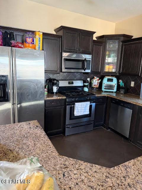 kitchen featuring decorative backsplash, appliances with stainless steel finishes, glass insert cabinets, dark brown cabinets, and light stone countertops