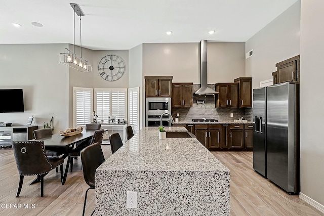 kitchen with tasteful backsplash, stainless steel appliances, wall chimney range hood, decorative light fixtures, and an island with sink