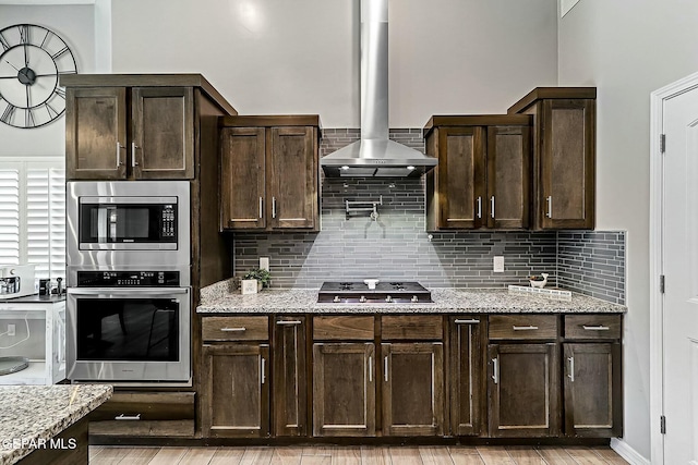 kitchen with wall chimney range hood, decorative backsplash, light stone counters, dark brown cabinetry, and stainless steel appliances