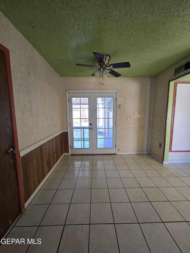 tiled empty room featuring french doors, a textured ceiling, ceiling fan, and wooden walls