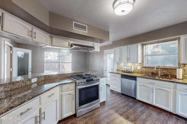 kitchen with wall chimney range hood, white cabinetry, appliances with stainless steel finishes, dark stone countertops, and sink