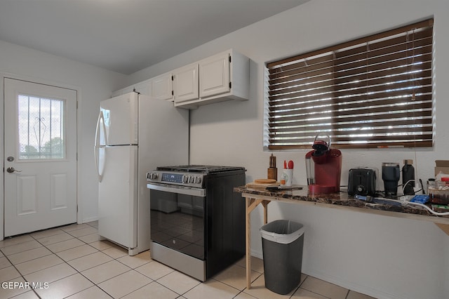 kitchen with stainless steel gas range oven, white refrigerator, light tile patterned floors, white cabinetry, and dark stone counters