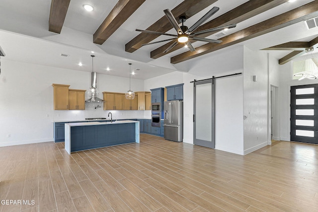 kitchen with appliances with stainless steel finishes, beamed ceiling, an island with sink, hanging light fixtures, and a barn door