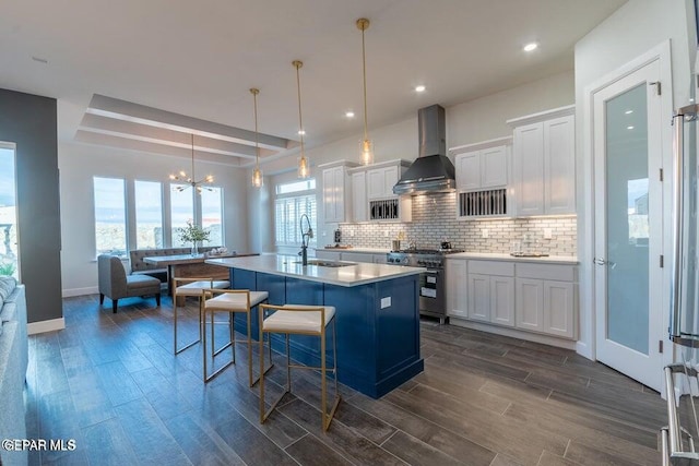 kitchen with wall chimney exhaust hood, pendant lighting, high end range, an island with sink, and white cabinetry