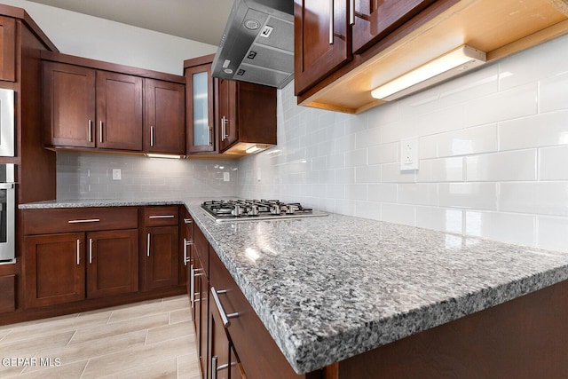 kitchen with decorative backsplash, wall chimney exhaust hood, glass insert cabinets, light stone countertops, and stainless steel gas cooktop