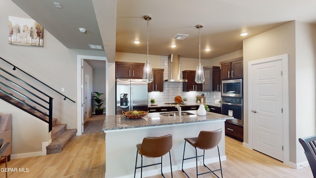 kitchen with sink, wall chimney exhaust hood, decorative backsplash, a kitchen island with sink, and appliances with stainless steel finishes