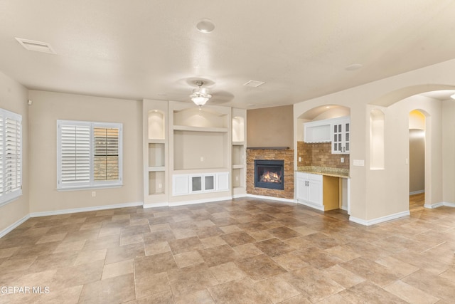 unfurnished living room featuring ceiling fan, built in features, and a stone fireplace