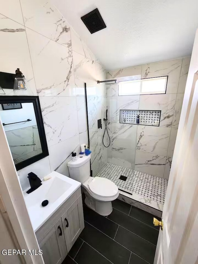 bathroom with tile walls, tiled shower, vanity, and toilet