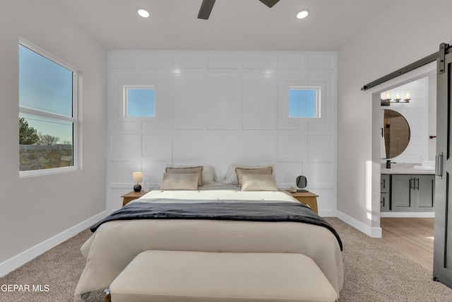 bedroom with a barn door, connected bathroom, light colored carpet, and ceiling fan