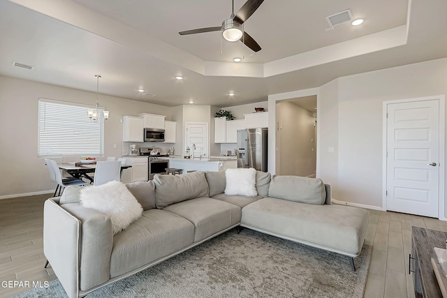 living room with sink, ceiling fan with notable chandelier, and a tray ceiling