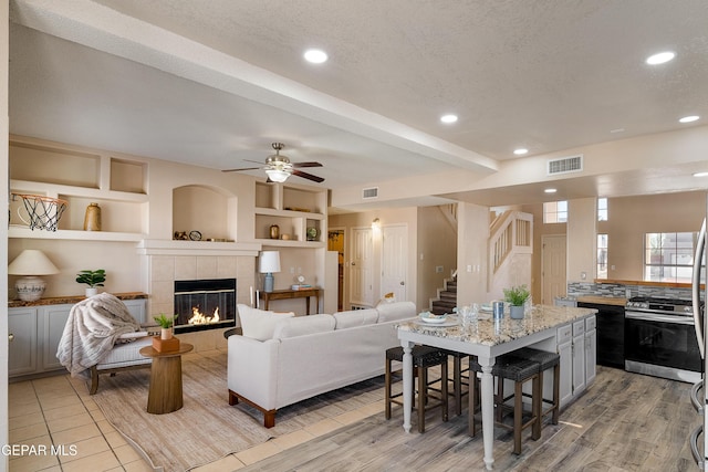 living room with ceiling fan, a tiled fireplace, built in features, a textured ceiling, and beam ceiling