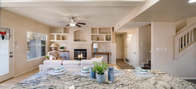 living room with ceiling fan, built in shelves, a tile fireplace, and a textured ceiling