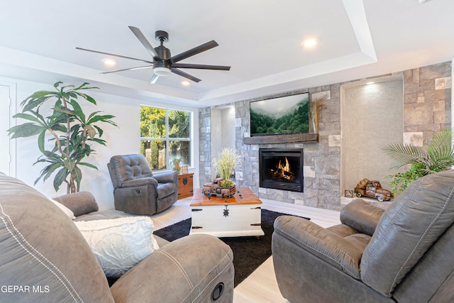 living room featuring ceiling fan, light wood-type flooring, a stone fireplace, and a tray ceiling