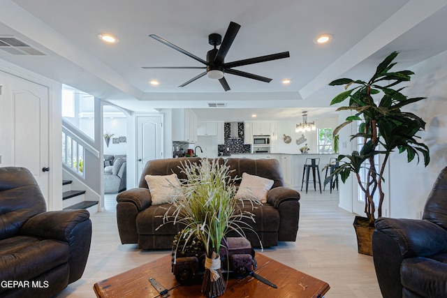 living room with light wood-type flooring, a tray ceiling, and ceiling fan with notable chandelier