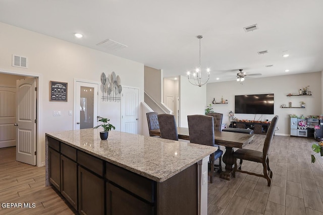 kitchen with dark brown cabinets, a kitchen island, pendant lighting, ceiling fan with notable chandelier, and light stone counters