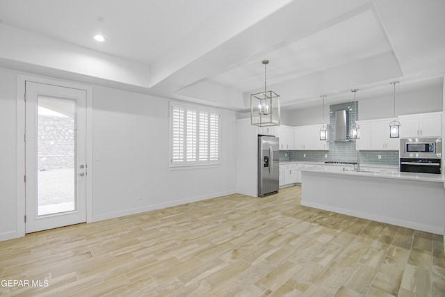 kitchen with appliances with stainless steel finishes, white cabinetry, hanging light fixtures, a tray ceiling, and wall chimney range hood