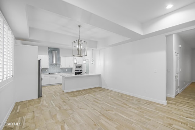 kitchen with wall chimney range hood, white cabinetry, stainless steel appliances, a tray ceiling, and decorative light fixtures