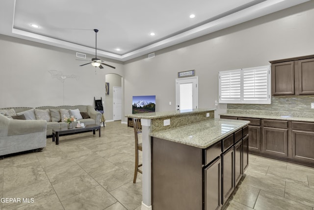 kitchen featuring a kitchen breakfast bar, ceiling fan, light stone countertops, a tray ceiling, and dark brown cabinetry