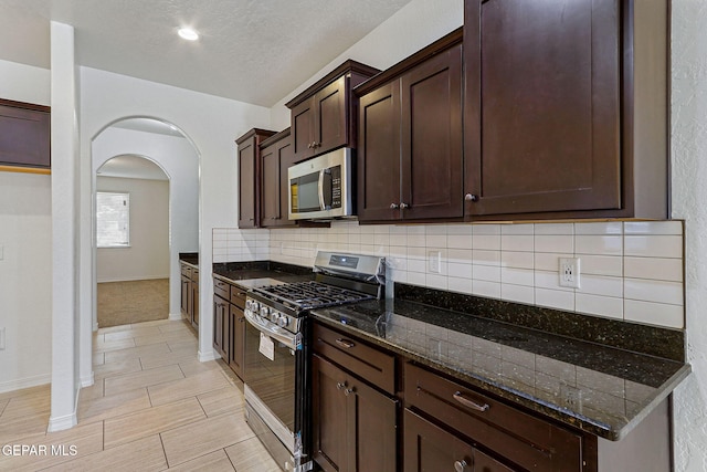 kitchen with a textured ceiling, dark brown cabinets, dark stone countertops, light colored carpet, and stainless steel appliances