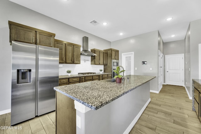 kitchen with wall chimney exhaust hood, light stone counters, a center island with sink, decorative backsplash, and stainless steel appliances