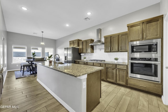 kitchen with light stone countertops, appliances with stainless steel finishes, wall chimney exhaust hood, hanging light fixtures, and a center island with sink