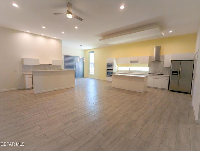 kitchen featuring wall chimney range hood, ceiling fan, appliances with stainless steel finishes, white cabinetry, and a kitchen island