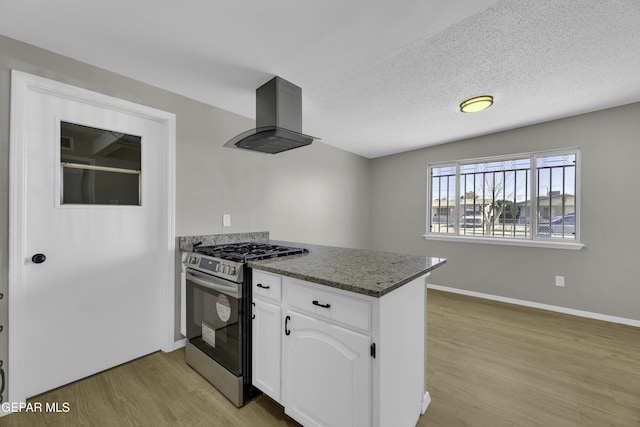 kitchen with stainless steel gas range, dark stone countertops, island range hood, white cabinets, and light wood-type flooring