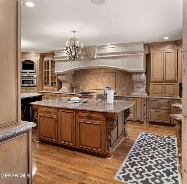 kitchen featuring light stone counters, pendant lighting, light wood finished floors, a kitchen island with sink, and stainless steel oven