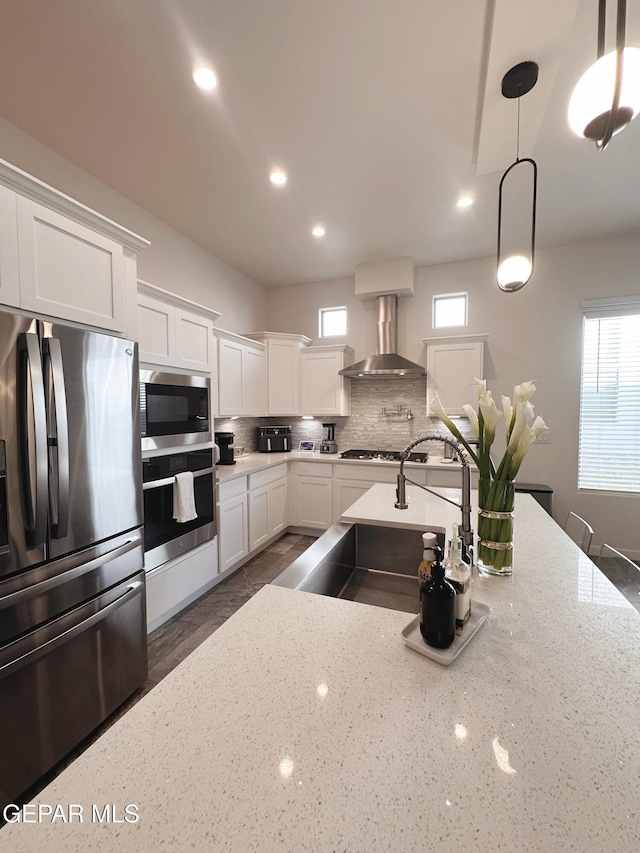 kitchen with appliances with stainless steel finishes, pendant lighting, light stone counters, and wall chimney exhaust hood