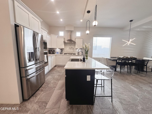 kitchen with white cabinets, wall chimney range hood, hanging light fixtures, stainless steel appliances, and a center island with sink