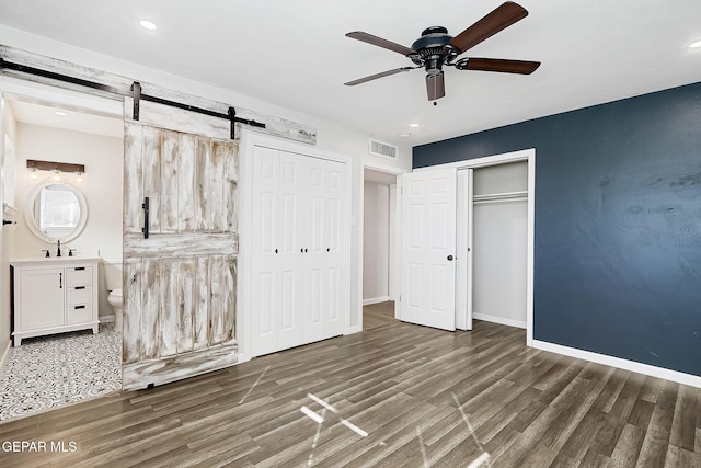 unfurnished bedroom featuring ensuite bath, dark wood-type flooring, a barn door, and ceiling fan