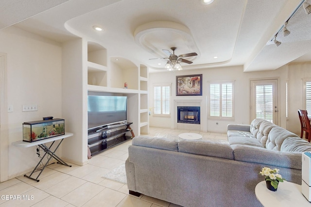 tiled living room featuring track lighting, a tray ceiling, built in features, ceiling fan, and a tiled fireplace