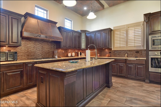 kitchen featuring premium range hood, stainless steel appliances, dark brown cabinetry, an island with sink, and decorative light fixtures