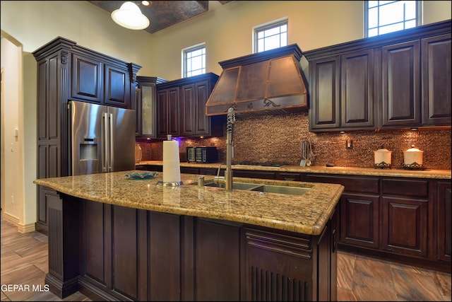 kitchen with custom exhaust hood, tasteful backsplash, stainless steel fridge with ice dispenser, dark brown cabinets, and a center island with sink