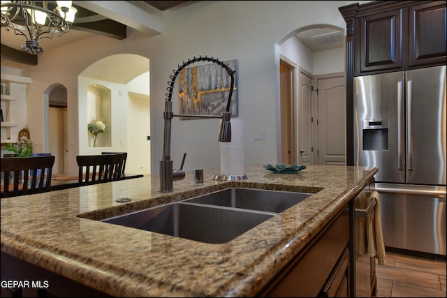 kitchen featuring sink, beam ceiling, dark brown cabinets, light stone counters, and stainless steel fridge with ice dispenser