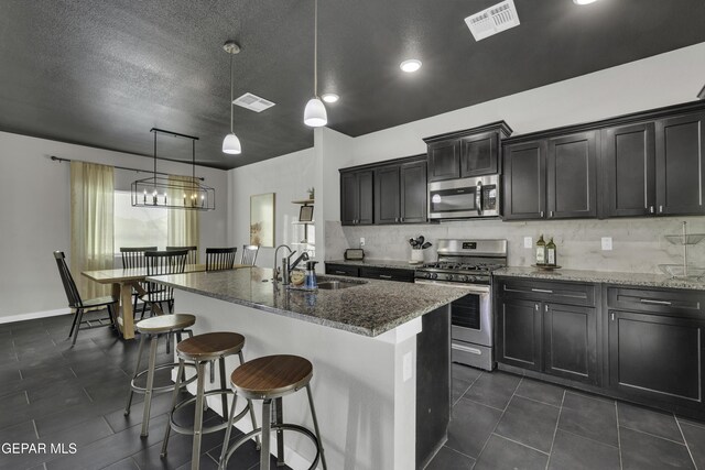 kitchen with sink, dark stone countertops, hanging light fixtures, stainless steel appliances, and a center island with sink