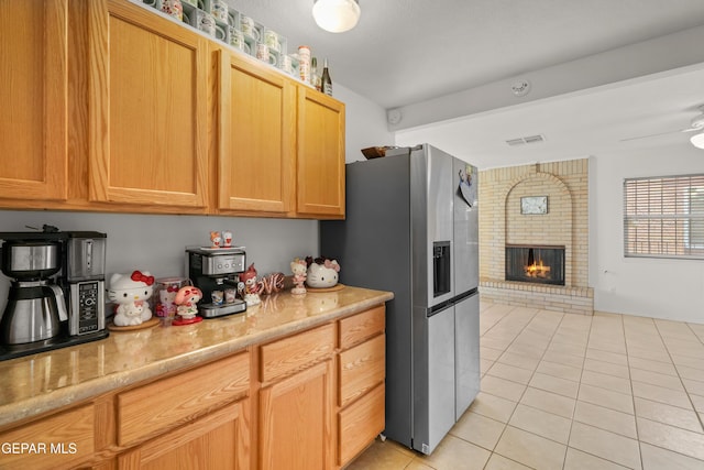 kitchen with stainless steel refrigerator with ice dispenser, light brown cabinetry, light tile patterned floors, ceiling fan, and a fireplace