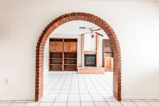 unfurnished living room with a brick fireplace, light tile patterned floors, ceiling fan, and built in shelves