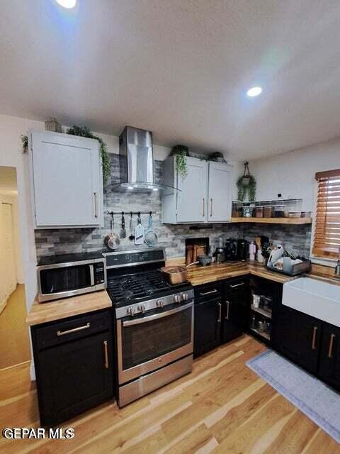 kitchen with light wood-type flooring, appliances with stainless steel finishes, white cabinets, wall chimney range hood, and backsplash