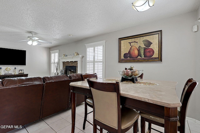 tiled dining space with ceiling fan, a stone fireplace, and a textured ceiling