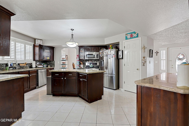 kitchen with dark brown cabinets, light tile patterned floors, pendant lighting, stainless steel appliances, and a kitchen island with sink